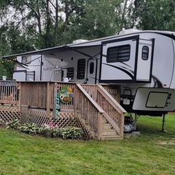 Large camper trailer with wooden deck and stairs in a green outdoor setting.