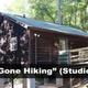 Exterior of a rustic studio cabin labeled 'Gone Hiking' showing porch and surrounding forest — representative on-property lodging image from the homepage.