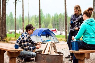 Tent campsite scene showing people around a fire ring with tents and surrounding trees — depicts tent camping facilities and atmosphere relevant to the property’s campsite offerings (included on the campground page).