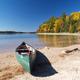 A green canoe rests on a sandy beach. Blue water and a wooded shoreline beginning to show fall color are in the distance.