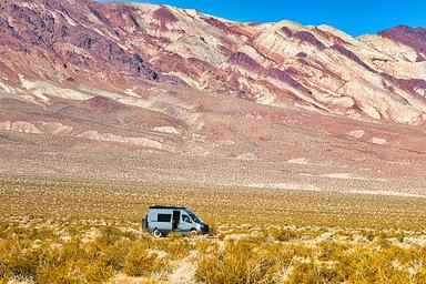 Gray campervan on roadside campsite with green brush vegetation in the foreground and dramatic reddish and white stratified mountains in the far background.