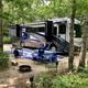 On-site RV site photo showing a Class A motorhome parked on a paved pitch with campsite furnishings and fire pit — clearly illustrates RV accommodations and site layout (from the site gallery).
