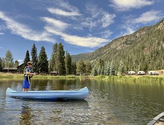 A person stands on a blue canoe, fishing on a calm lake surrounded by pine trees, mountains, and a few lakeside cabins under a blue sky.