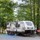 Shows an RV parked at a gravel, wooded campsite with outdoor setup — clear depiction of RV site layout and utilities. Matches on‑site 'RV Sites' imagery in the property gallery.