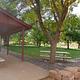 A large tree is in front of a white picnic table. A brown pavilion is to the left, a white picnic table is underneath it and the picnic table is on top of concrete. A grassy area with many trees are in the background. 