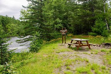 A gravel and grassy campsite to the right of a river. The campsite has a wooden picnic table, a brown wooden sign, and a trail that leads into the woods with tall dense green trees.
