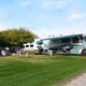RVs parked in a campground with trees and a grassy area, one person sitting at a picnic table, and a clear blue sky.