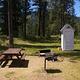 The picnic table, fire ring and pedestal grill are behind the cabin partially shaded by pines and green pastures. 