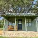 Green cabin with a covered front porch, a wooden bench and a stone foundation beneath a large oak tree