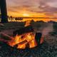 Foreground fire pit with logs on a lakeshore at sunset; visible dock and boat silhouettes in background — a clear on-site campground activity/amenity image taken from the property's homepage photo gallery.