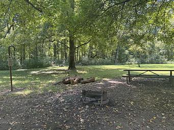 A brown picnic table sits to the right of a campsite. A campfire ring and lantern post can be seen in the foreground.