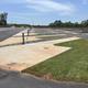 Empty parking lot with newly paved walkways, green grass, and rocks under a partly cloudy sky.