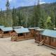 A row of cozy wooden cabins with green metal roofs sits in a forest clearing, mountains and tall trees in the background, under a blue sky.