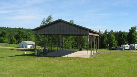 Open-sided pavilion/covered activity area on lawn with RVs and utility buildings in background — useful facility image showing common area (homepage image).