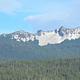 Pine trees in silhouette on near shore, sunny blue lake, and conifer covered hills below a rocky ridge line and partly cloudy sky.