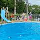 Four people jumping into a blue swimming pool on a sunny day, with a slide visible.