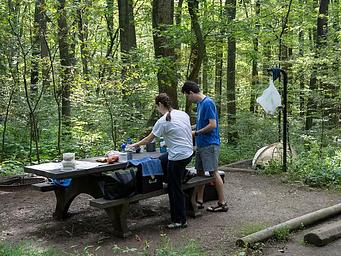 Man and woman standing next to picnic table on wood chip campsite.  On the tent pad behind the campers is a tent.  Waste back hangs from lantern pole.  Site is surrounded by dense forest.