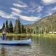 A person stands on a blue canoe, fishing on a calm lake surrounded by pine trees, mountains, and a few lakeside cabins under a blue sky.
