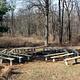 Outdoor fire pit with 18 wooden benches on concrete posts in circular pattern.  Center of circle has a metal fire ring/grill. Winter photo, surrounding trees  have no leaves.