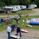 Overhead-ish view of a grassy activity area with cornhole courts, inflatable game, and RVs in the background — shows campground recreation space and site layout.