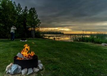Campfire in a ring on grassy lakeshore with person photographing the sunset — shows campsite atmosphere and firepit amenities by the lake (homepage gallery image).