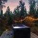 An outdoor hot tub on a wooden deck in a forest, illuminated by string lights hanging from trees at dusk.