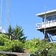 Lookout building on ridge with Forest Service i.d. sign, communications tower, green shrubs and conifer trees backed by blue sky.