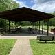 A view of an open sided pavilion with picnic tables, pedestal BBQ grill, and benches with a background of green vegetation. 