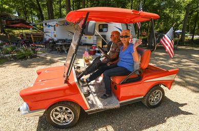 Guests in a decorated golf cart with RVs and wooded campsites in the background — highlights guest experience and campground roadways/parking areas (from site gallery).