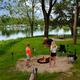 Lakeside tenting/firepit scene with people and grassy shoreline — demonstrates tent sites and lake access, consistent with the site’s tenting/seasonal images.