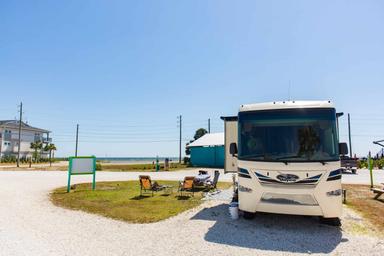 A white RV is parked at a campground near the beach with two empty chairs and a small table set up outside on a sunny day.