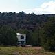 A large motor home is parked in a camp site. There are trees lined on both sides and a hillside with red rocks and trees can be seen from the site.