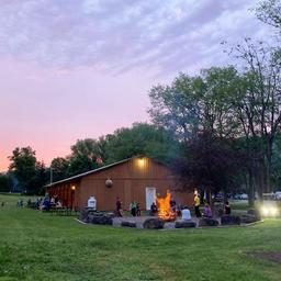 Wide outdoor shot at dusk showing a lodge/building, grassy lawn and a central campfire ring with people — good view of campground social area and facilities.