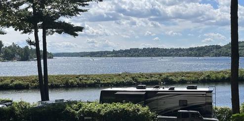 Scenic lake view with RV and greenery under a partly cloudy sky.