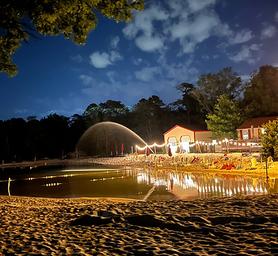 Clean spring-fed lake at evening at Sleepy Hollow Campground with activity center, chairs, and lights in background.