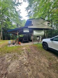 RV under a shelter in a wooded campsite with a white vehicle parked nearby.