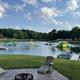 Wide-shot of the spring-fed lake with inflatable water park features and dock, with surrounding campsites visible — strong overview of the signature lake attraction.