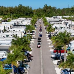 High-angle view looking straight down a main park street lined with RVs and palm trees; highlights site spacing and community events (file labeled bayfest-parade on the property site).