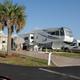 A large RV beside palm trees, with chairs, an umbrella, and people enjoying the outdoor space.