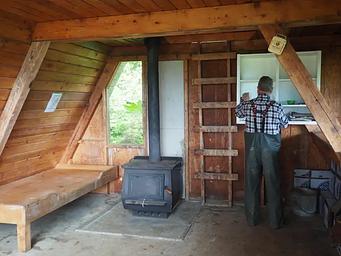 Man at shelves inside wood cabin with black stove and wood bunk