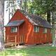 Small, orange-red cabin with ramp to front door in sun dappled conifer forest.