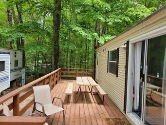 Wooden deck beside a beige cabin, with picnic table, chair, and wooded forest view.