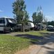Wide exterior photo showing a row of RVs parked along paved road with grassy sites and trees — clear overall view of the campground’s RV sites; file appears on the property front page as a site/gallery image.