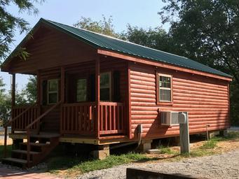 Stand-alone cabin on gravel pad with porch and window AC unit — depicts on-site cabin accommodations (from 'ways to stay' photos on the campground page).