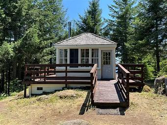 Short ramp leading onto wrap around porch of white cabin, fir trees surround the cabin.