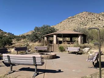 Uncovered fire pit surrounded by benches, with the covered pavilion with picnic table in the background.