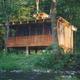 Wooden cabin on stilts in a wooded area, with a porch and small chimney.