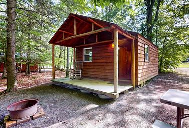 Close photo of a rustic cabin with porch, wooded setting and fire ring — useful to illustrate cabin accommodations and woodsy campsite atmosphere from the site’s Cabins section.