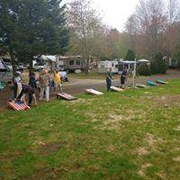 Open grassy area with RVs in background and cornhole game in foreground — shows recreational activity space and campsite arrangement; from the About Us page.