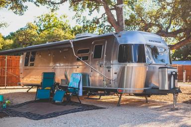 Airstream-style RV parked on a gravel site with shade and outdoor chairs — shows RV pitch accommodations and site setup; sourced from the property gallery.